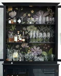 a black cabinet with floral wallpaper and wine glasses on the top shelf is filled with liquor