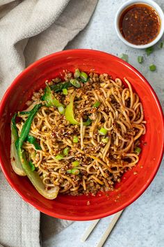 a red bowl filled with noodles and vegetables on top of a white table next to sauce