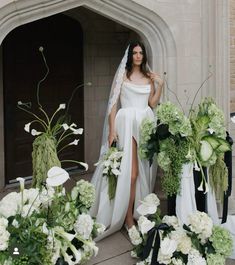 a woman in a wedding dress standing next to flowers and greenery on the ground