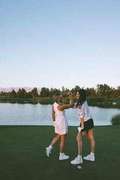 two women standing on top of a lush green field next to a body of water