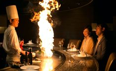 three people sitting at a table with food and wine in front of a fire pit