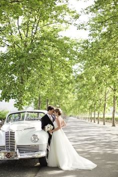 a bride and groom standing in front of an old car on the road near trees