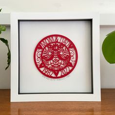 a red paper cut in a white frame on a wooden table next to a green plant