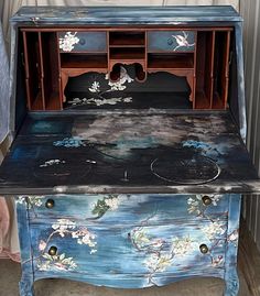 an old desk painted blue with flowers and birds on the top, sitting in front of a wall