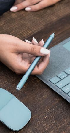 a person is holding a pencil near a keyboard and mouse on a wooden table with a laptop in the background