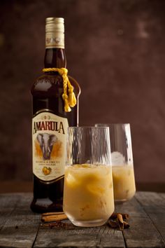 two glasses filled with liquid next to a bottle and cinnamon sticks on a wooden table