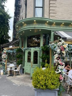 people sitting at tables in front of a building with flowers on the outside and green doors