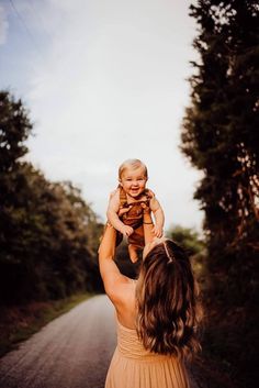 a woman holding a baby up in the air while walking down a road with trees