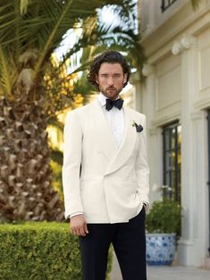 a man in a white tuxedo and bow tie standing on a sidewalk next to a palm tree