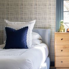 a bed with white sheets and blue pillows next to a wooden dresser in front of a window