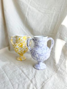 two white and yellow vases sitting on top of a cloth covered tableclothed surface