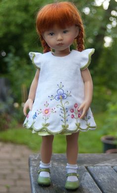 a doll with red hair wearing a white dress and green shoes standing on a wooden table