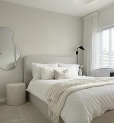 a bedroom with white bedding, pillows and a round mirror on the wall above it