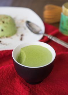 a bowl of green soup on top of a red table cloth next to a spoon