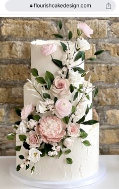 a white wedding cake with pink flowers and greenery on the top is displayed in front of a brick wall