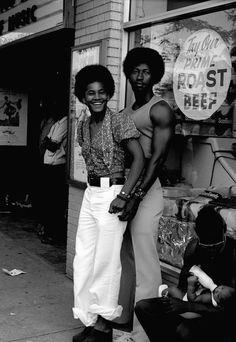 two people standing on the sidewalk in front of a store, one man is wearing white pants