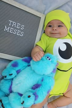 a baby in a green shirt is holding a blue stuffed animal next to a sign that says tres mess