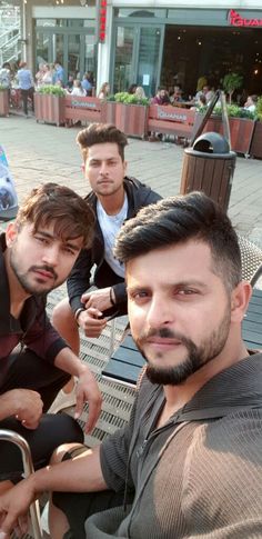 three men are sitting on a bench in front of an outdoor seating area with tables and chairs