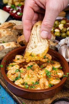 a hand is dipping some bread into a bowl of shrimp and olives with breadcrumbs on the side