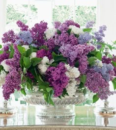 a glass vase filled with purple and white flowers on top of a table next to a window