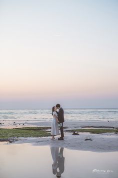 a bride and groom standing on the beach at sunset with their reflection in the wet sand