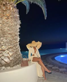 a woman sitting on a wall next to a palm tree at night with her legs crossed