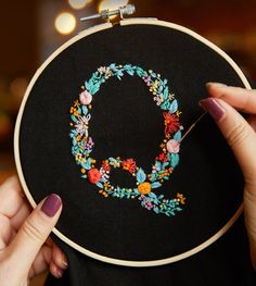 a woman holding up a black embroidery hoop with a floral letter q on it and a needle in the middle