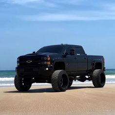 a large black truck parked on top of a sandy beach next to the ocean and blue sky