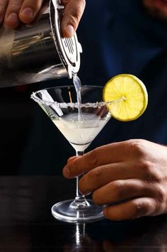 someone pouring a martini into a glass with a lemon wedge in it and another person's hand on the side