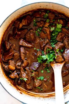 a pot filled with meat and garnish on top of a table next to a wooden spoon