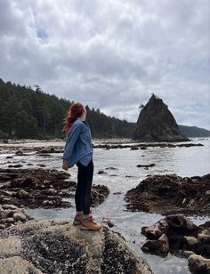 a woman standing on top of a rocky beach next to the ocean with trees in the background