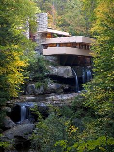 a house in the woods surrounded by trees and rocks with a waterfall running through it