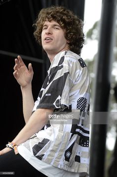 a young man with curly hair on stage at an outdoor music festival, holding his hands up