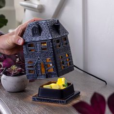 a person holding a house shaped candle holder on a table next to a potted plant