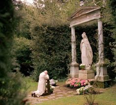 a man kneeling down next to a statue in a garden with flowers and trees around him