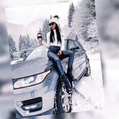 two people sitting on the hood of a car in the snow with trees behind them