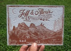 a person holding up a business card with mountains in the background and grass behind it