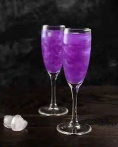 two wine glasses filled with purple liquid next to an ice cube on a wooden table