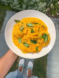 a person holding a plate of pasta with basil leaves on it in front of their feet