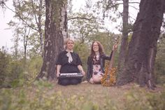 two women sitting on the ground in front of trees and holding musical instruments, both smiling at the camera