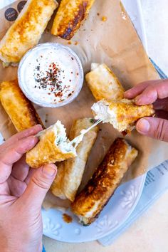 two hands holding food over a plate with dipping sauce