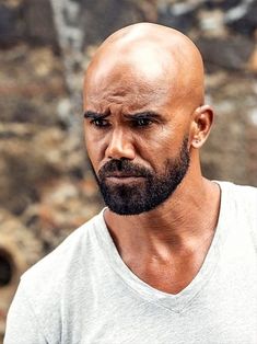 a man with a bald head wearing a white shirt and looking at the camera while standing in front of a stone wall