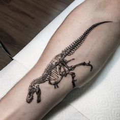 a black and white image of a dinosaur skeleton tattoo on the left arm, with a lizard in it's claws