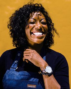 a smiling woman with an apron and watch on her wrist, standing in front of a yellow wall