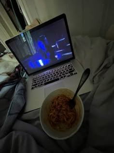 an open laptop computer sitting on top of a bed next to a bowl of noodles