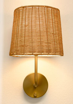 a wicker lamp on a white wall with a gold base and light bulb in the corner