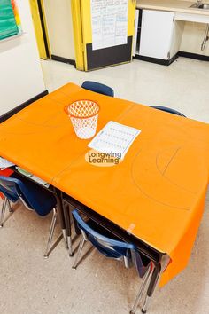 an orange table with blue chairs and a basket on it in the middle of a classroom