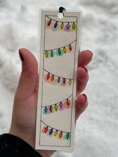 a hand holding a card with christmas lights on it in the middle of snow covered ground