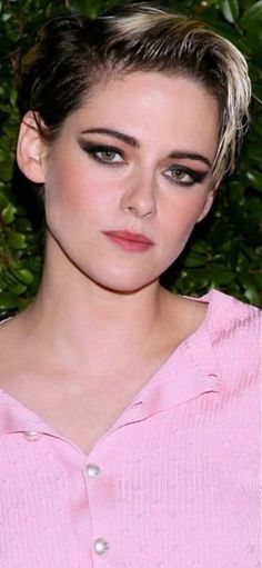 a close up of a person wearing a pink shirt and black eyeliners with green leaves in the background