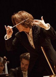 conductor in black suit and white shirt with orchestra behind him, holding his hand up to the side